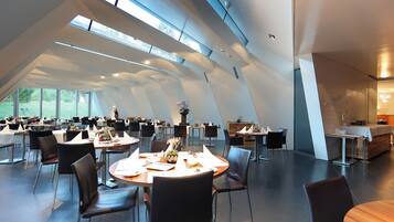 A modern dining area with multiple tables and chairs, large windows, and a high ceiling.