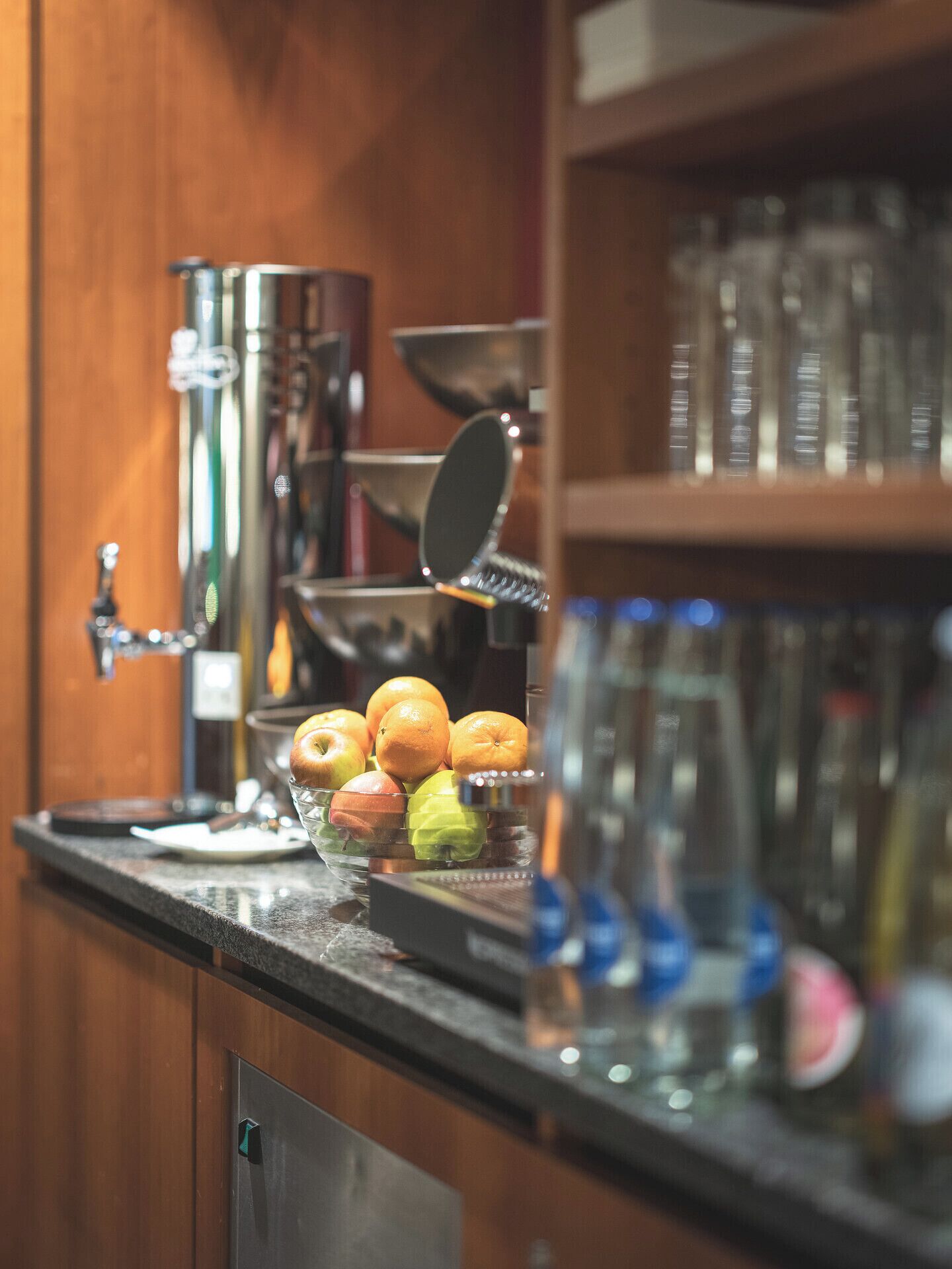Eine Küchenarbeitsplatte mit Obstschale, Äpfeln und Orangen, einer Kaffeemaschine und einem Glas Wasser.