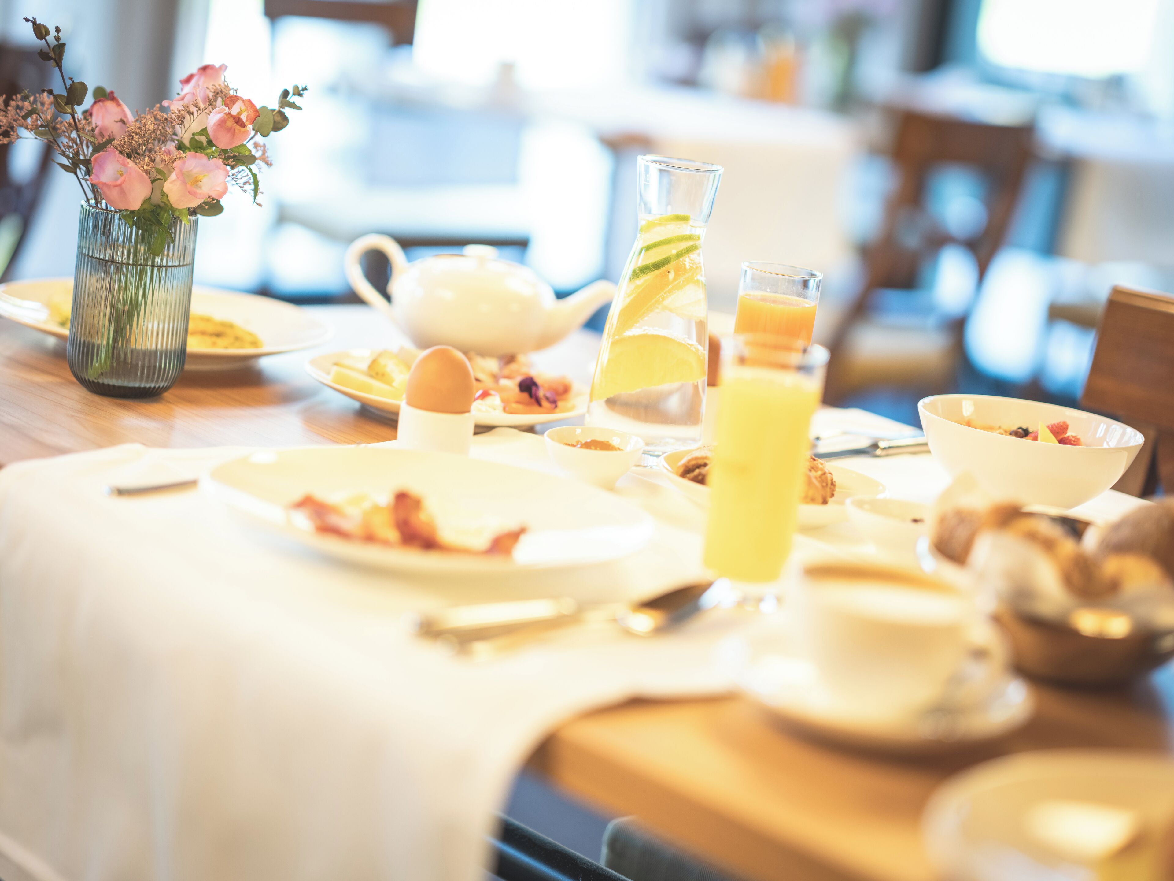 Una mesa de desayuno con platos, una jarrón, un vaso de jugo de naranja y un florero con flores.