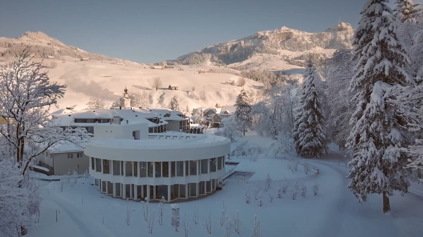 A snow-covered building with a circular layout, surrounded by snow-laden trees and mountains in the background.