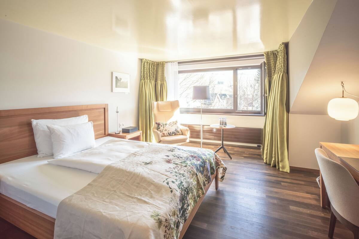 Une chambre d’hôtel avec un grand lit, un bureau, une chaise, une petite table et une fenêtre avec des rideaux.