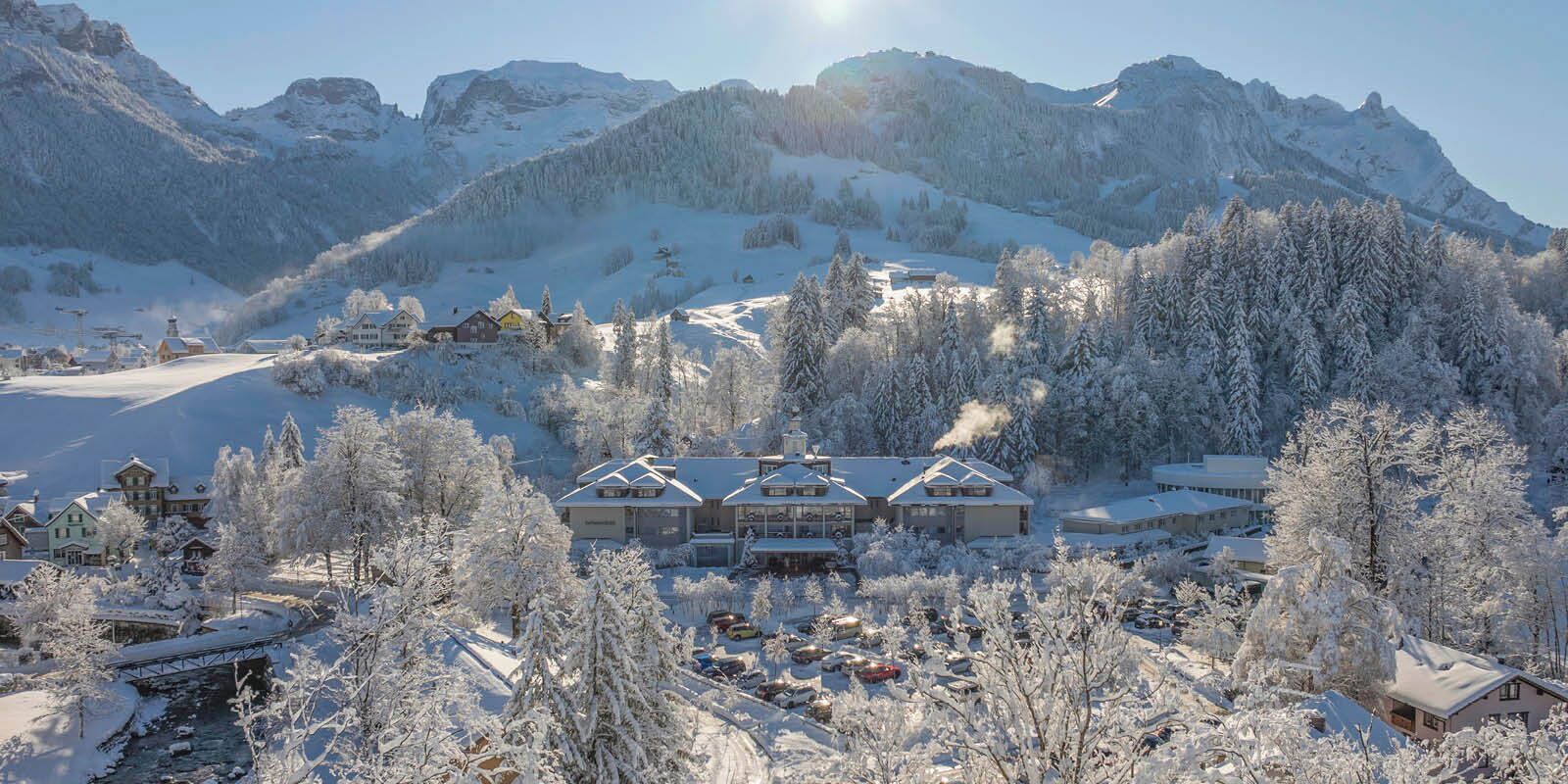 A snow-covered mountain resort with multiple buildings, a bridge, and a parking area.