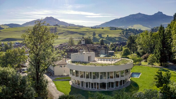 Un edificio moderno con diseño circular, rodeado de abundante vegetación y un paisaje montañoso.
