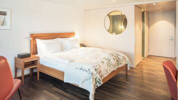 Una habitación de hotel moderna con una cama de madera, una mesita de noche con un teléfono, una silla roja y un espejo redondo en la pared.