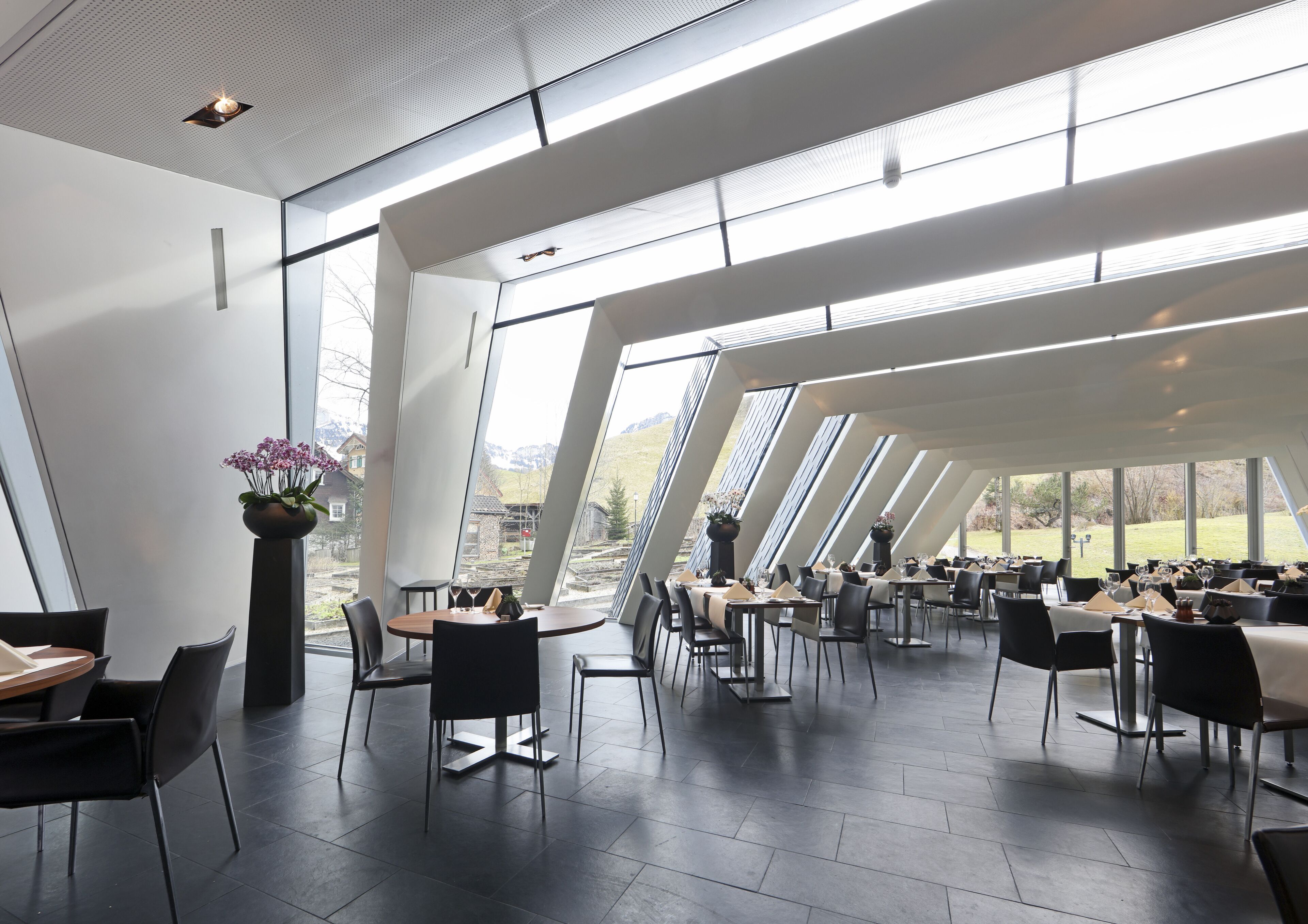 Un restaurante moderno con amplios ventanales y un techo alto. Las mesas están dispuestas para comer, y se ven sillas y mesas.