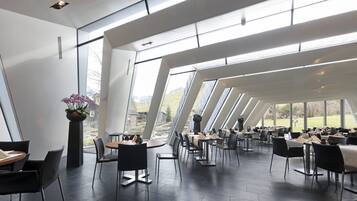 Un restaurante moderno con amplios ventanales y un techo alto. Las mesas están dispuestas para comer, y se ven sillas y mesas.