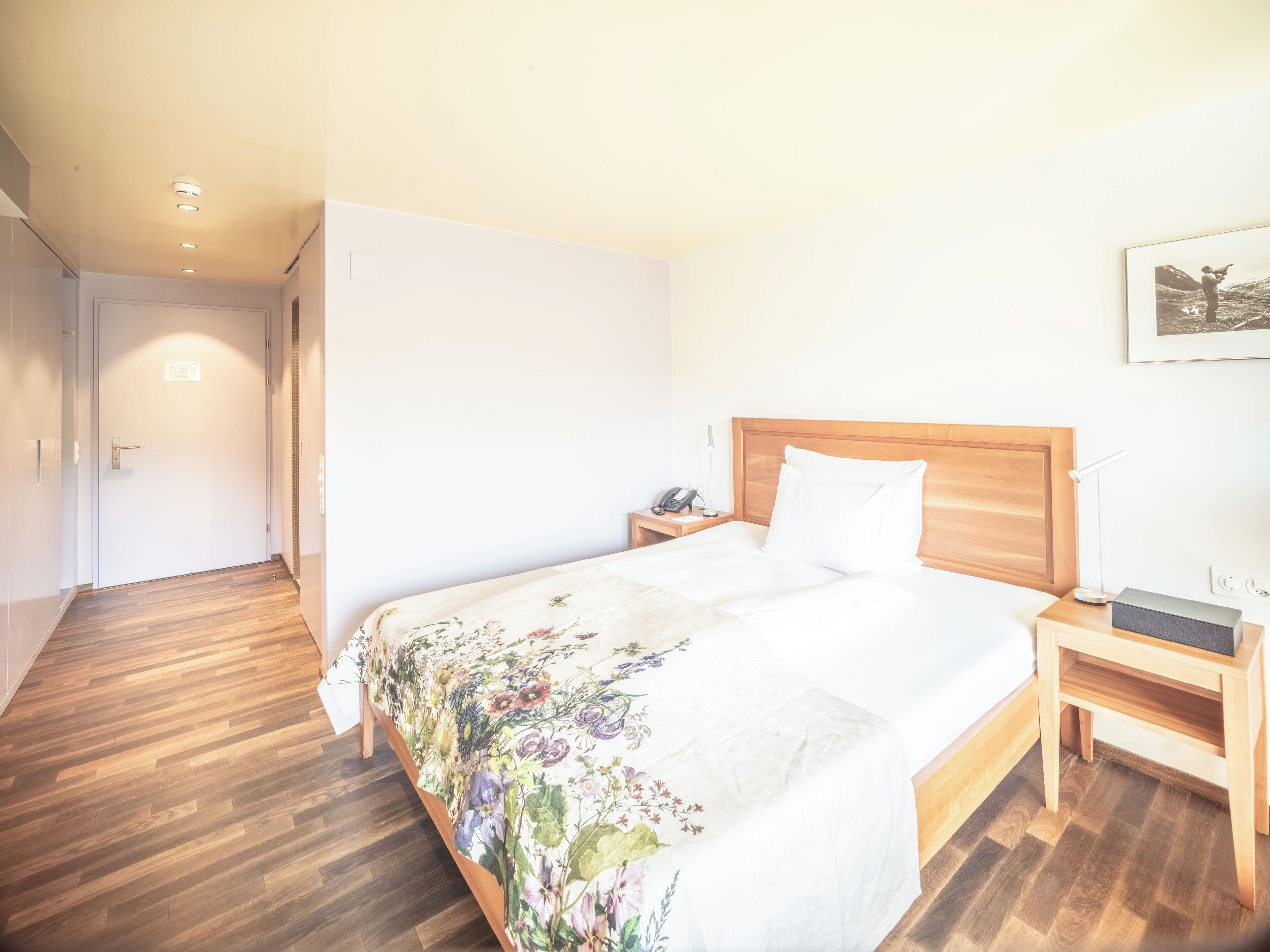 Ein Hotelzimmer mit Holzboden, einem Bett mit floralem Überwurf, einem Holz-Kopfteil, einem Nachttisch mit Lampe, einem Telefon und einem gerahmten Bild an der Wand.