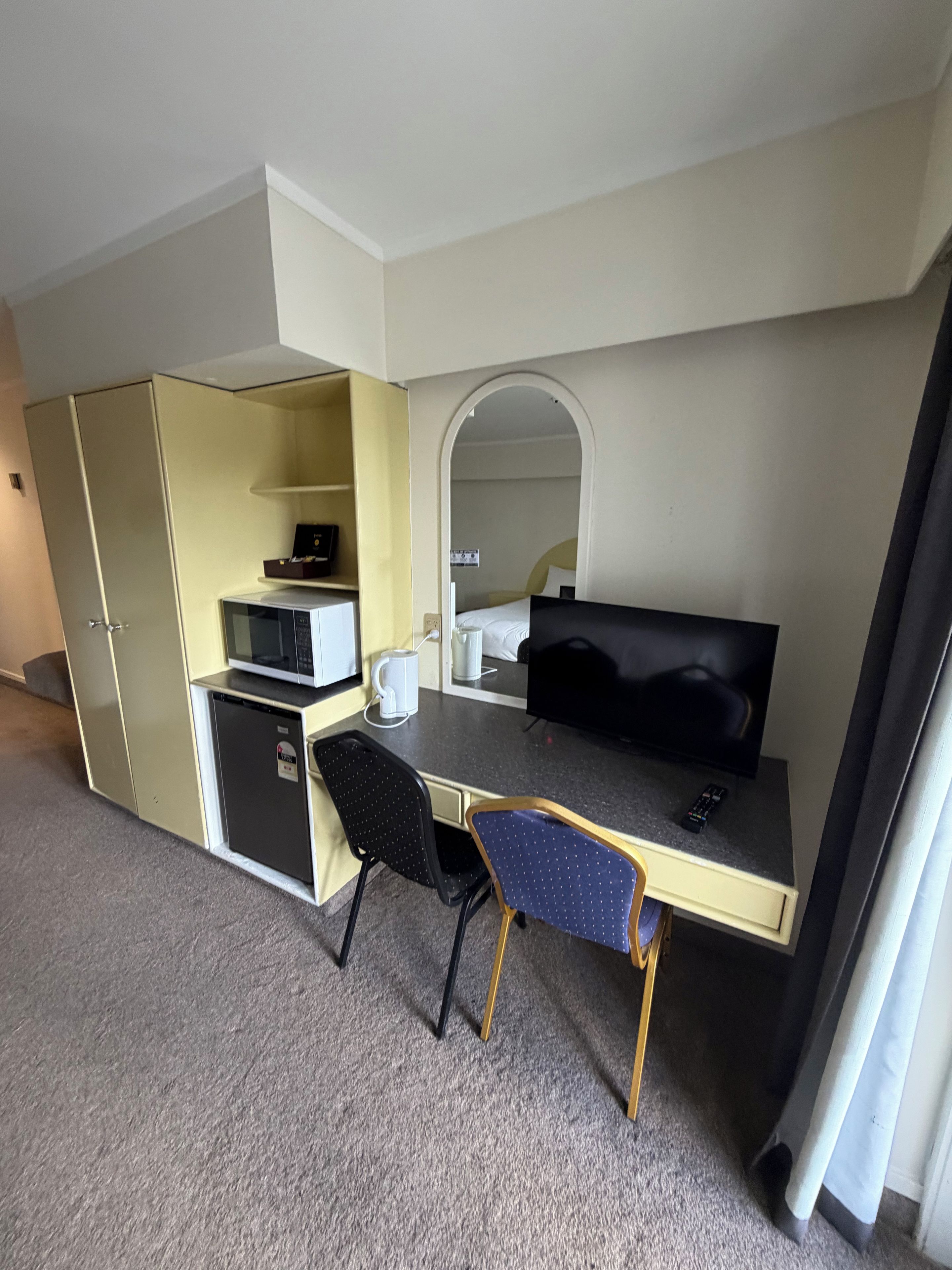 Et lille hotelværelse med tekøkken, en enkeltseng, et skrivebord med stol og et fjernsyn.