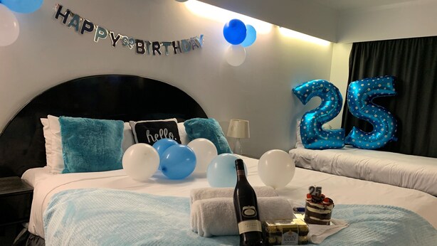 Et hotelværelse med seng, skilt med ”Tillykke med fødselsdagen”, balloner og en ballonskulptur med tallet 25.