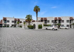 Un edificio residencial de varios pisos con un patio empedrado, coches aparcados y palmeras.