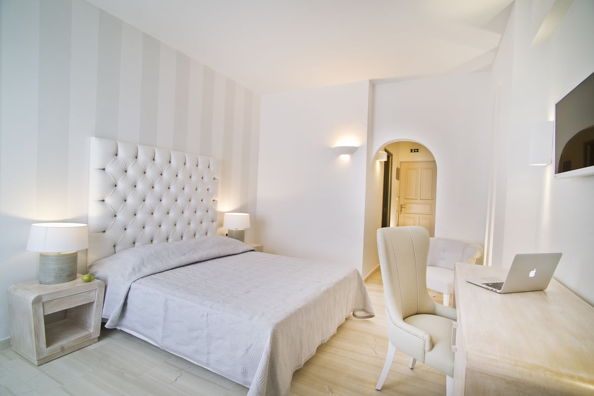 Un dormitorio moderno con cabecera capitonada, una mesita de noche con lámpara, una silla y un escritorio con computadora portátil.