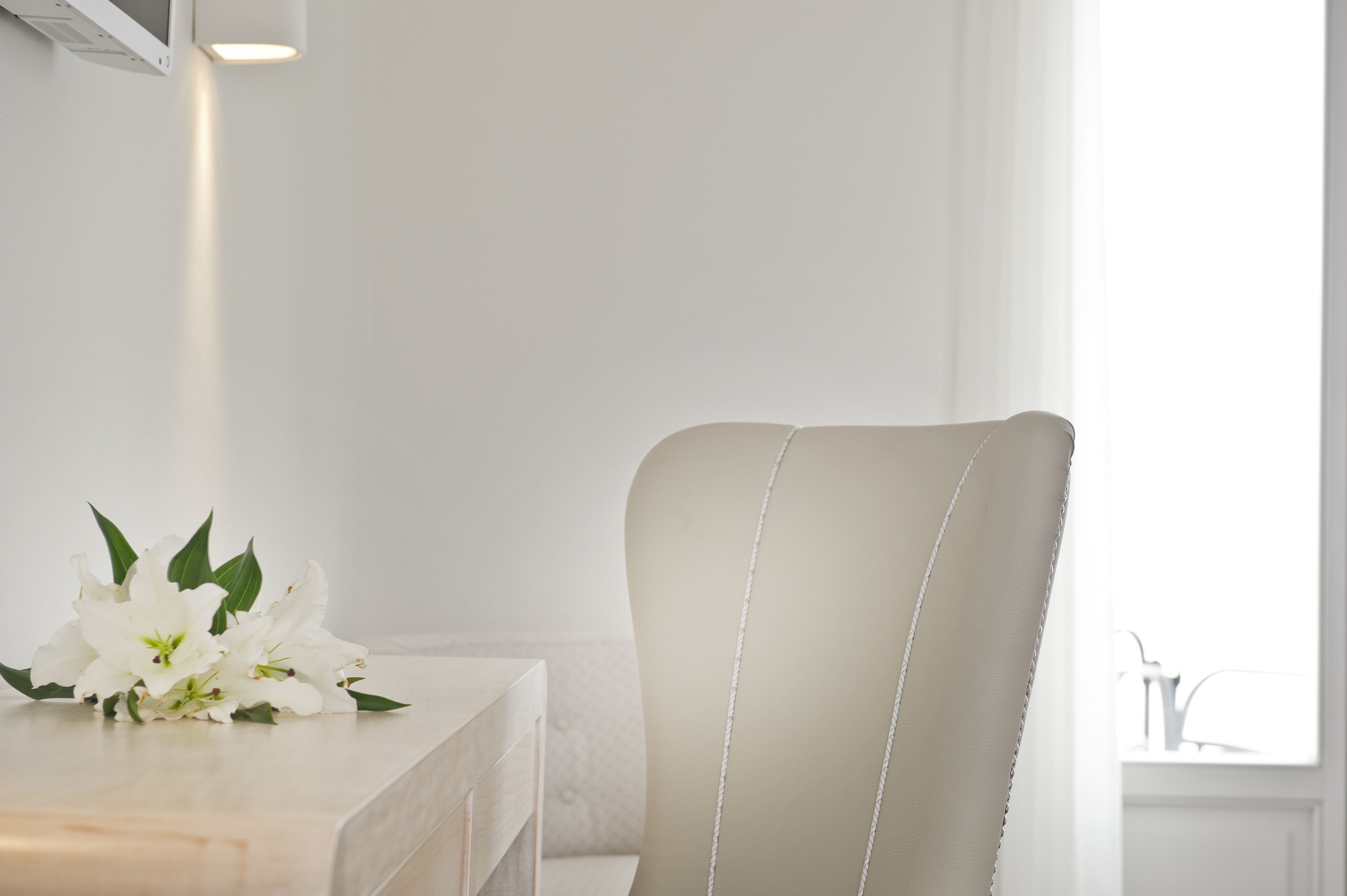Une pièce minimaliste avec une chaise blanche, une petite table recouverte d’une nappe blanche et une fenêtre laissant entrer la lumière naturelle.