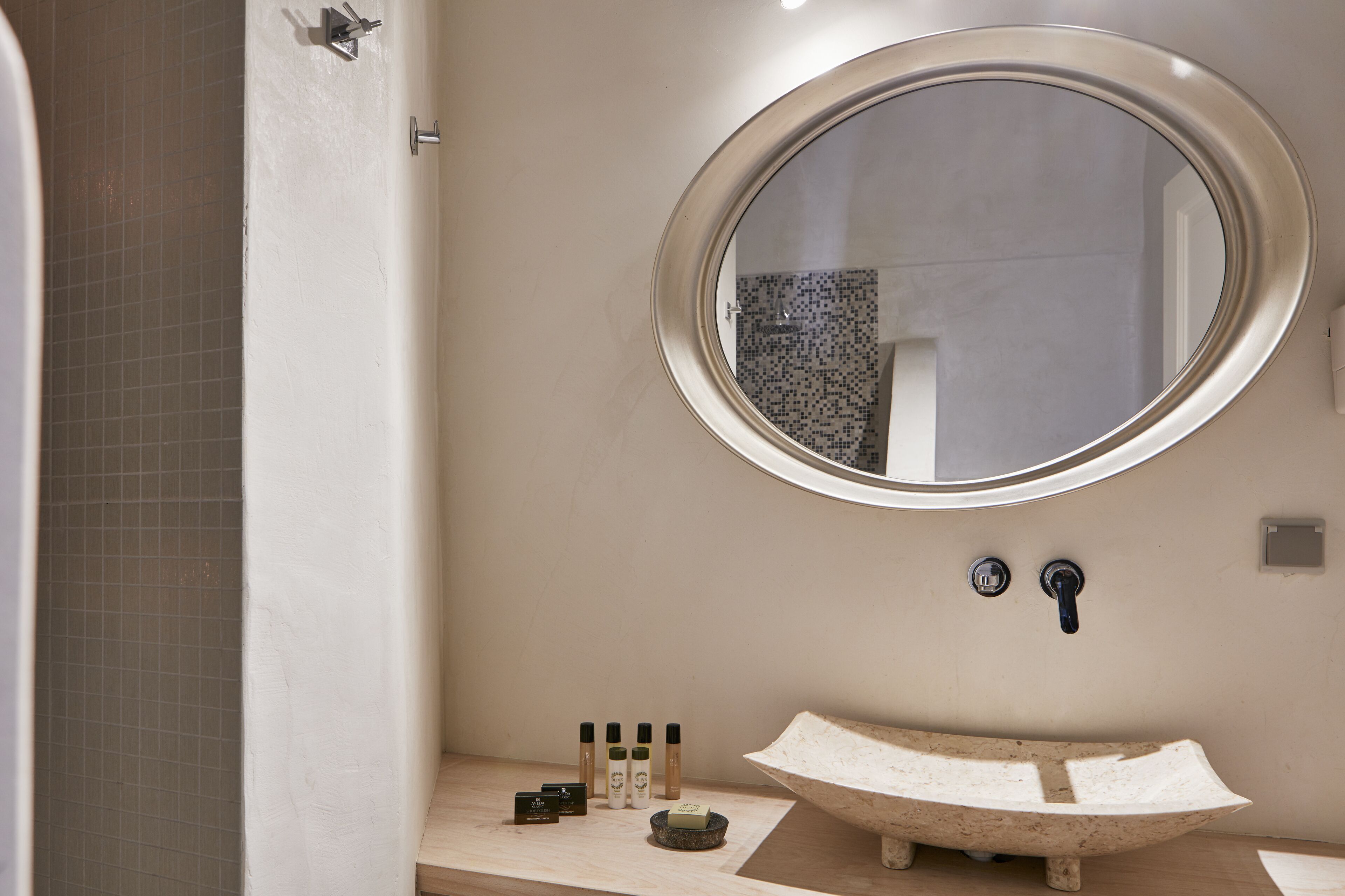 A bathroom with a round mirror, a stone sink, and toiletries on the countertop.