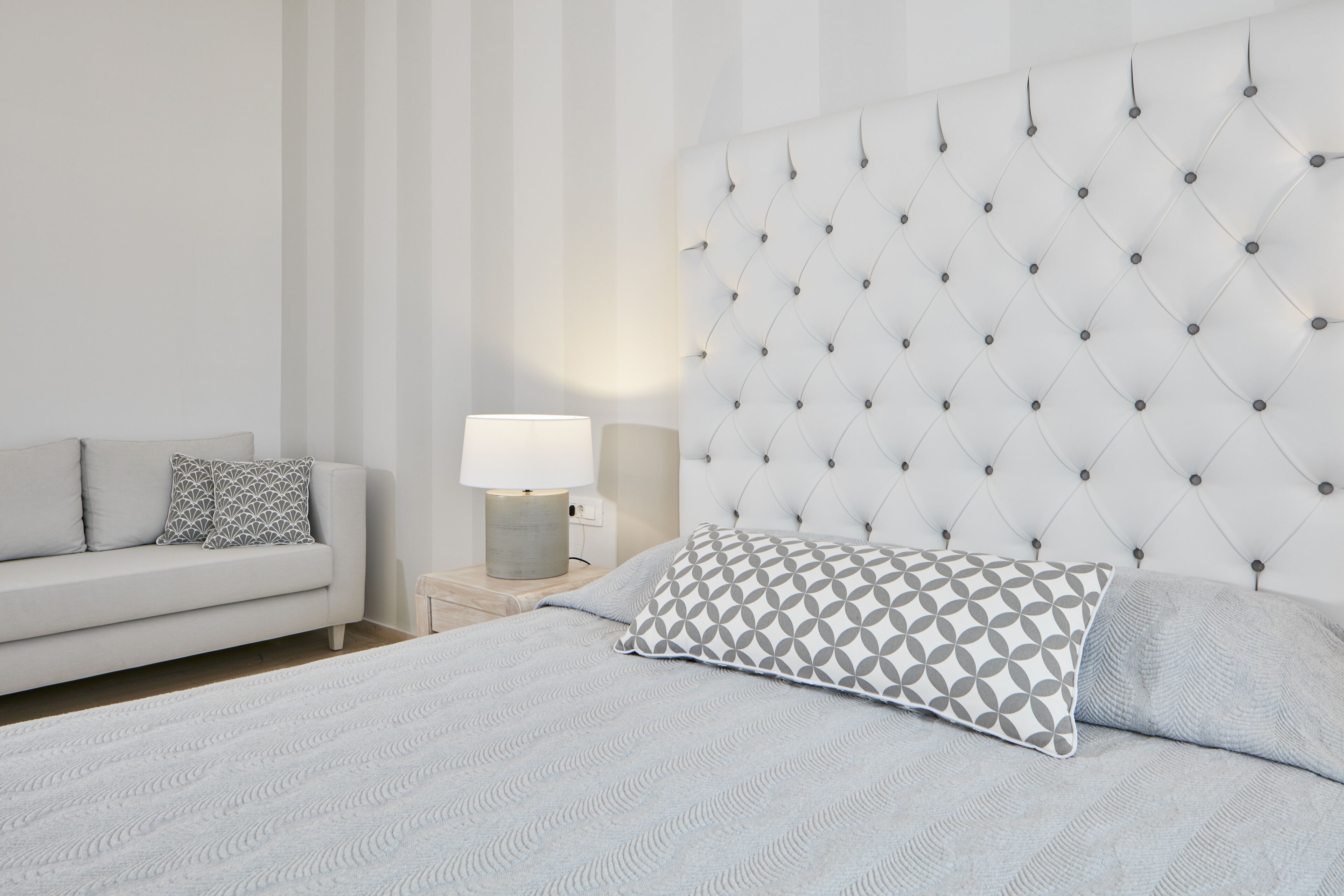 A bedroom with a tufted headboard, a bed with patterned pillows, a bedside table with a lamp, and a sofa with cushions.