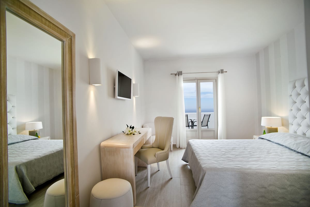 Habitación de hotel con cama, escritorio, silla, televisión y balcón con vistas.