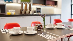 A breakfast table set with white cups, saucers, and a glass of orange juice. There are red chairs around the table, and a vase with purple flowers on the table.