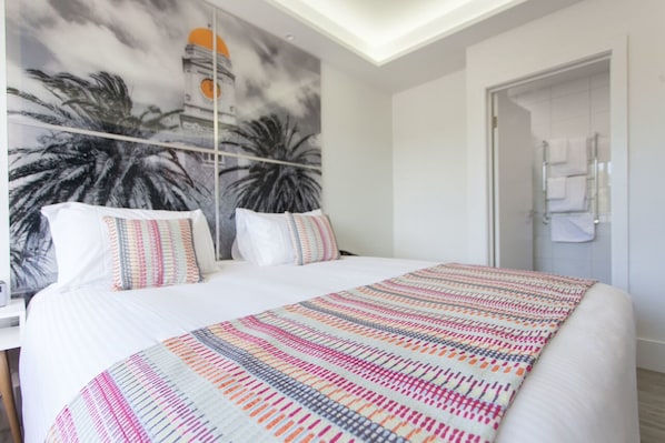 A bedroom with a large bed, a colorful striped blanket, and a mural of palm trees and a building on the wall.