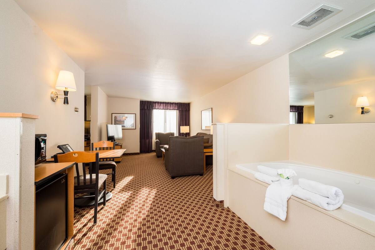 A hotel room with a desk, chairs, a sofa, a large mirror, and a bathtub with towels.