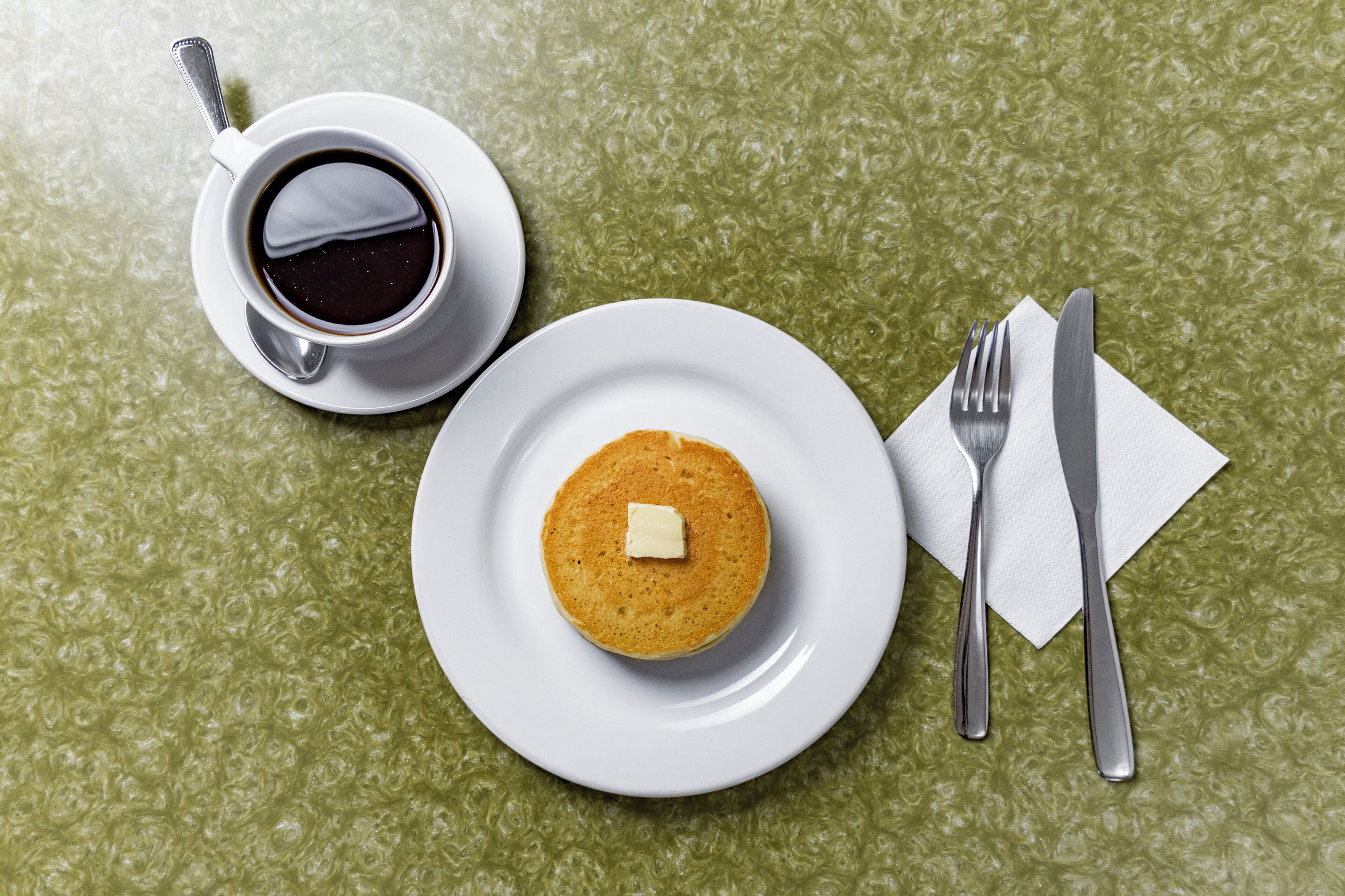 A breakfast setting with a plate of pancakes, a cup of coffee, and cutlery on a textured green table.