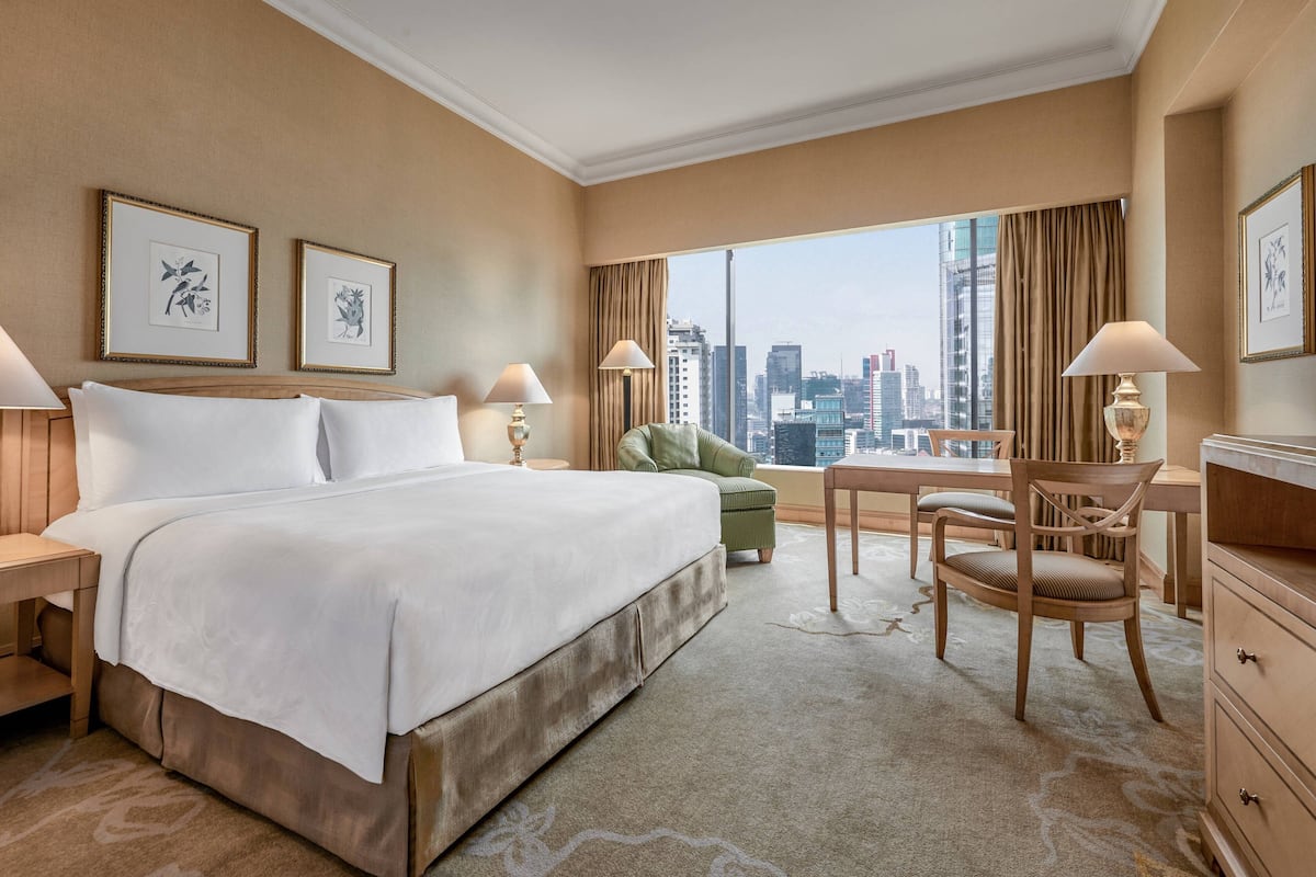 Ein Hotelzimmer mit einem großen Bett, zwei Nachttischen, einem Schreibtisch mit Stuhl und Blick auf die Stadt, sowie gerahmten Kunstwerken an den Wänden.