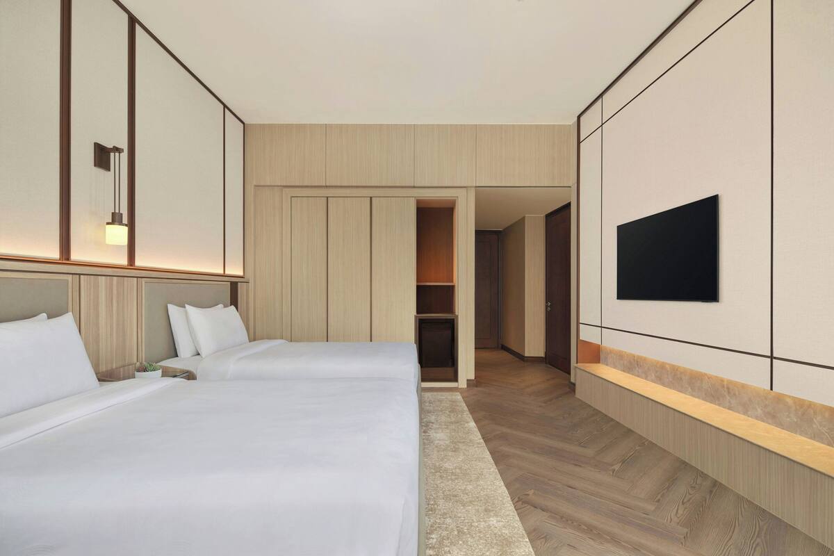 Ein Hotelzimmer mit zwei Betten, einem an der Wand befestigten Fernseher und einem zur Garderobe führenden Gang.