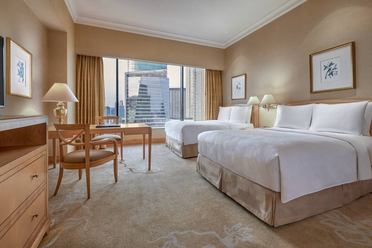 Ein Hotelzimmer mit zwei Betten, einem Schreibtisch, einem Stuhl und großen Fenstern mit Blick auf die Stadt.