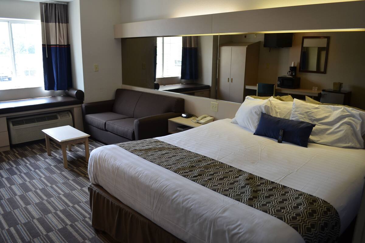 A hotel room with a large bed, a desk with a chair, a small table, a TV, and a mirror.