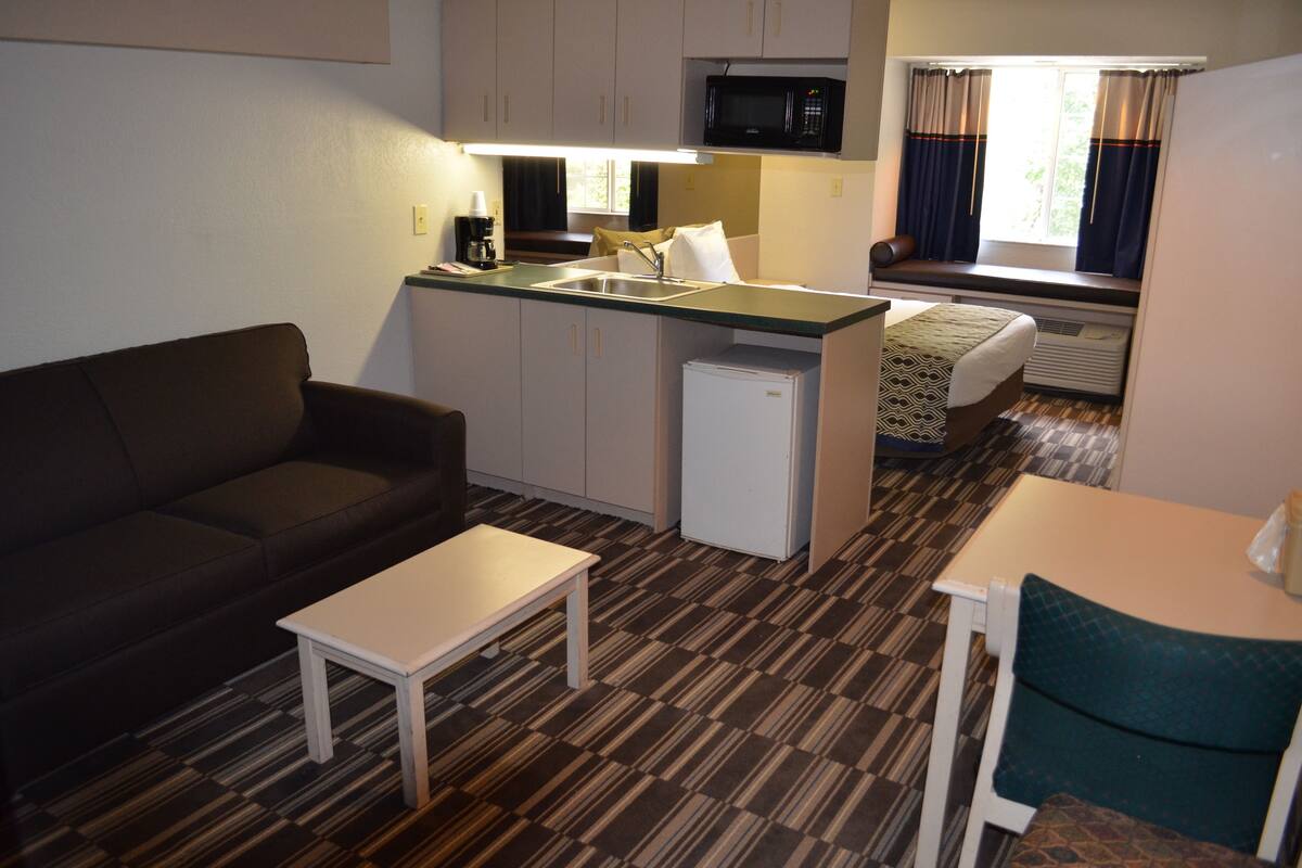 A compact room with a kitchenette, a sofa, a small table, and a window with curtains.