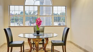 A dining area with a round glass table, two chairs, a vase with flowers, and a large arched window with a view of parked cars and buildings. - Stay Express Inn & Suites Atlanta (Union City)