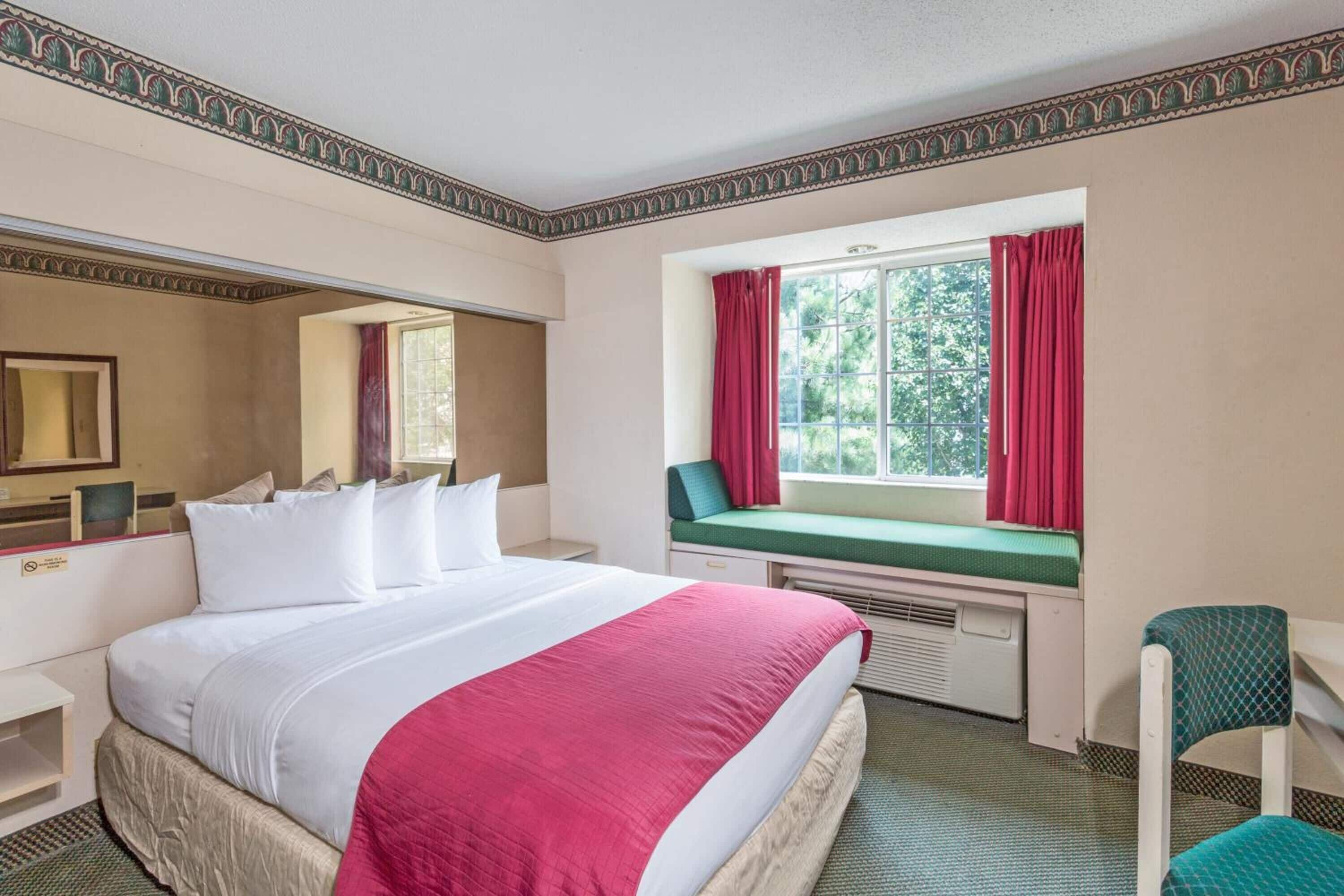 A hotel room with a large bed, a window with red curtains, a small table, and a chair.