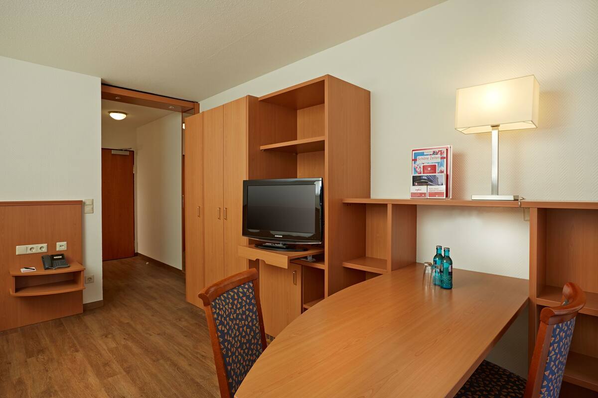 Une chambre d’hôtel avec une table en bois, des chaises, un téléviseur, une lampe et un mini-bar.