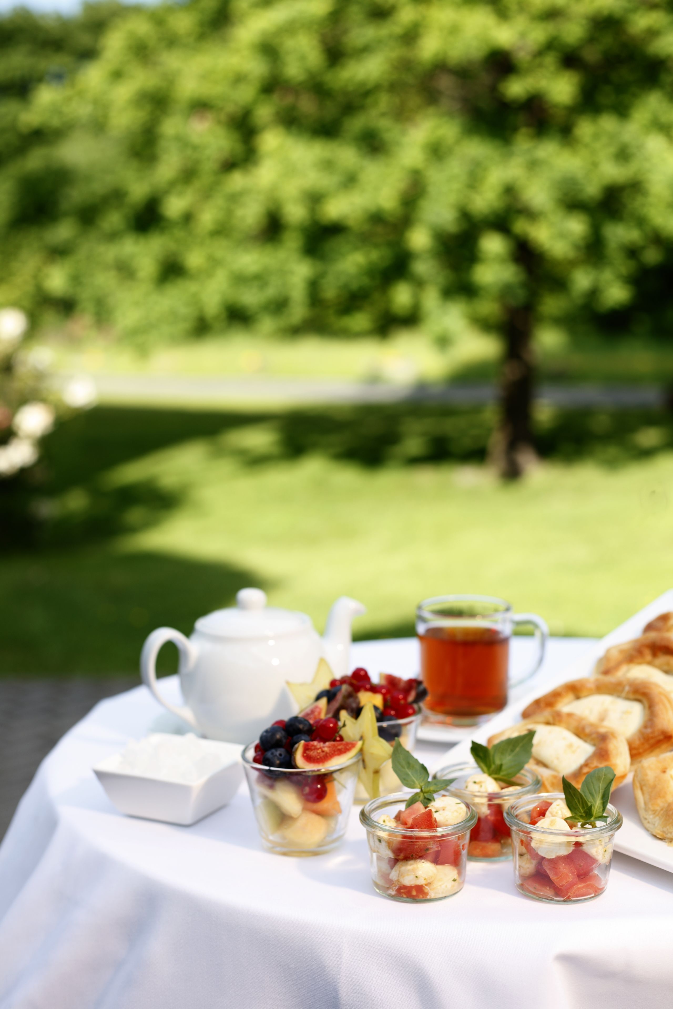 A table with a white tablecloth, a teapot, a glass of tea, a plate of pastries, and small glass cups with fruit and cream.