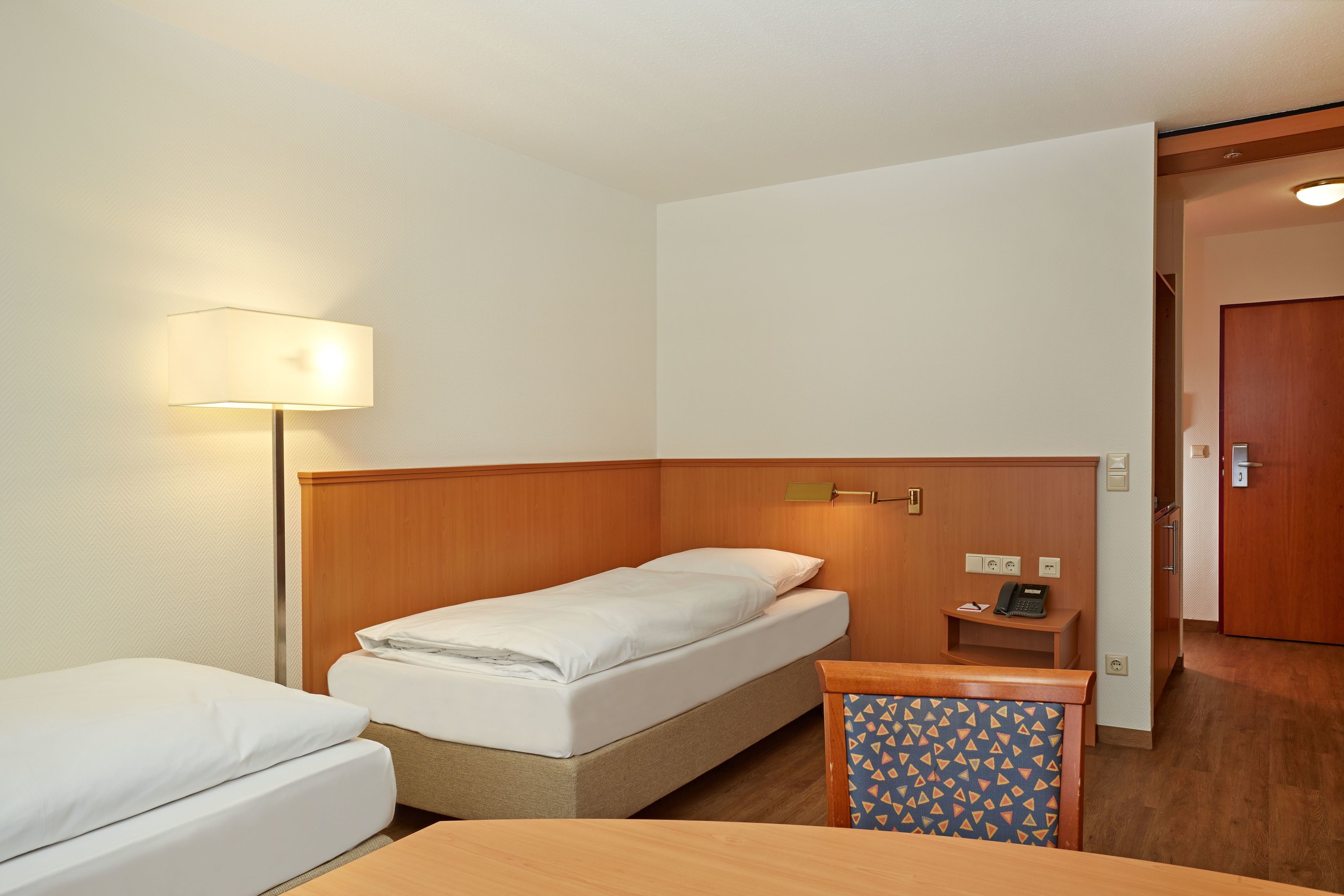 A hotel room with a single bed, a bedside table with a lamp, a small desk, and a chair with a patterned cushion.