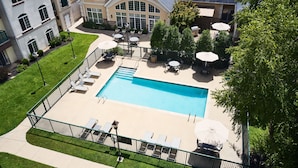 A rectangular pool with lounge chairs and umbrellas, surrounded by a fence and greenery.