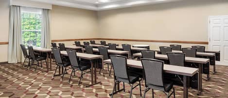 Un salón de conferencias con varias mesas y sillas, piso alfombrado y un amplio ventanal con cortinas.
