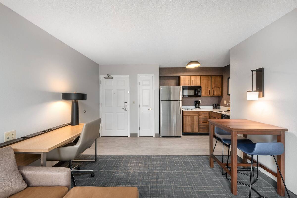 A compact hotel room with a kitchenette, a dining area with a table and chairs, a sofa, and a desk with a lamp.