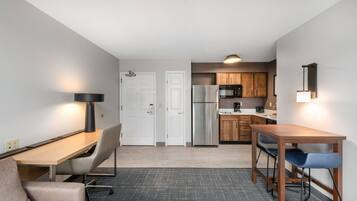 A compact hotel room with a kitchenette, a dining area with a table and chairs, a sofa, and a desk with a lamp.