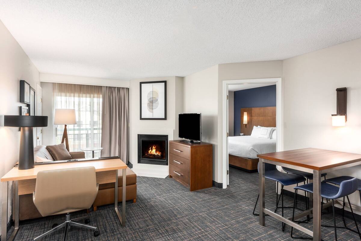 A hotel room with a desk, chair, TV, fireplace, bed, and a small dining area.