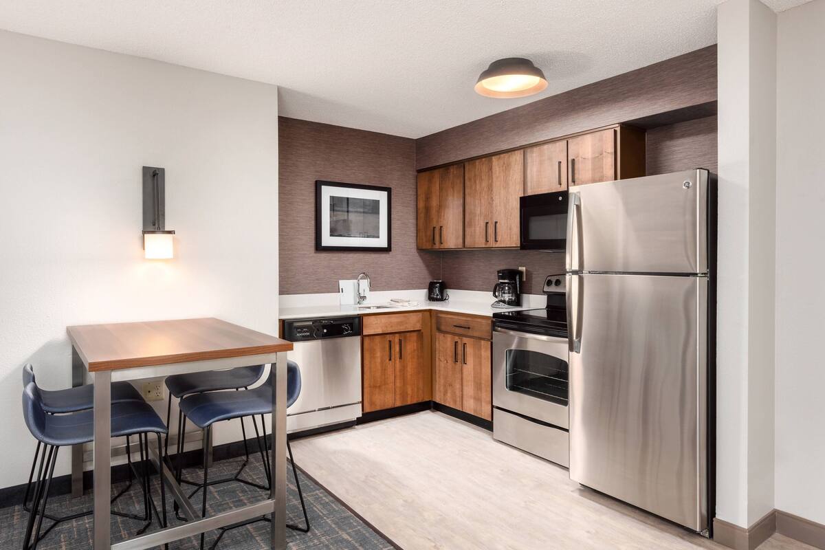 A compact kitchen with wooden cabinets, a stainless steel refrigerator, a dishwasher, and a coffee maker.