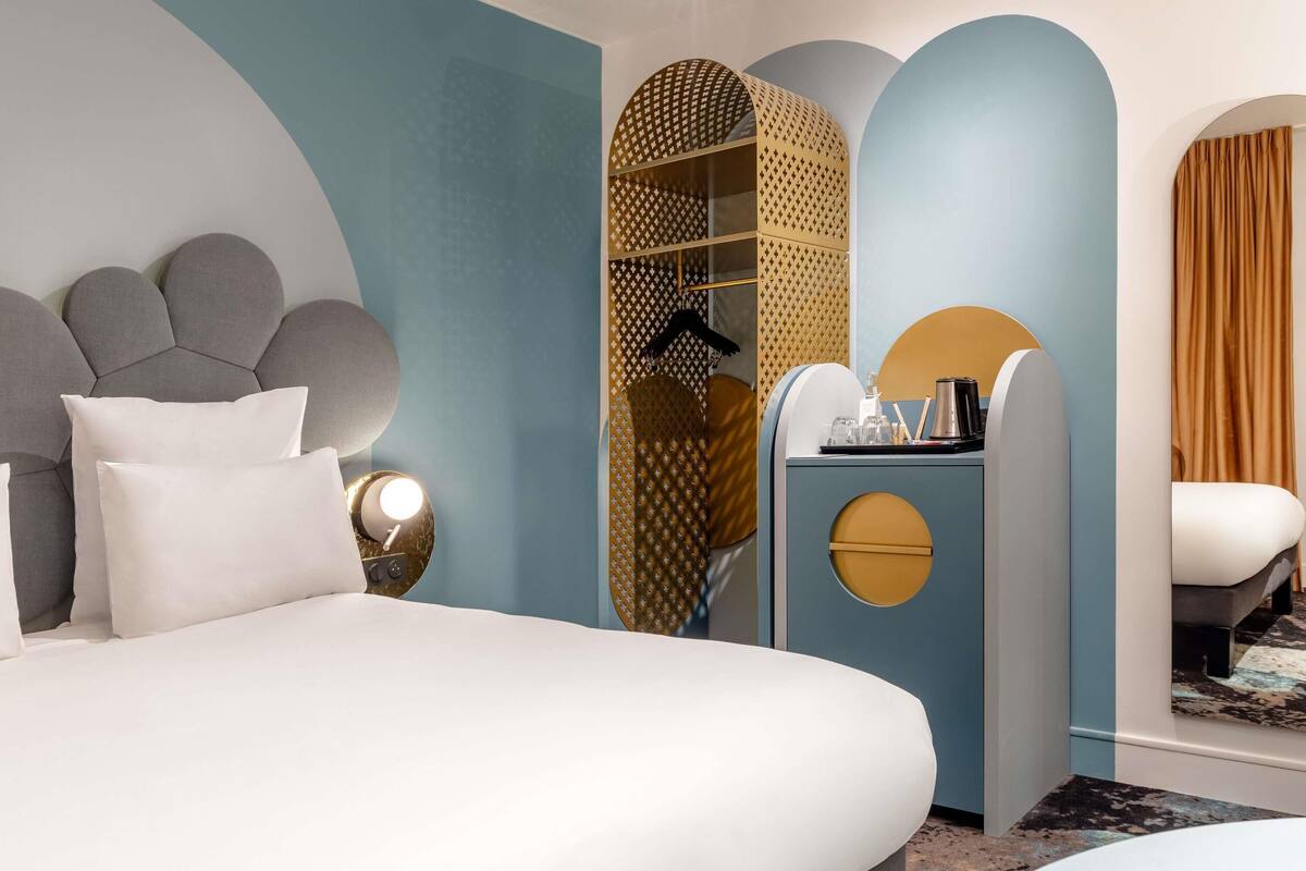 A modern hotel room with a large bed, a bedside table with a lamp, a wardrobe with patterned doors, and a seating area with a chair and a small table.