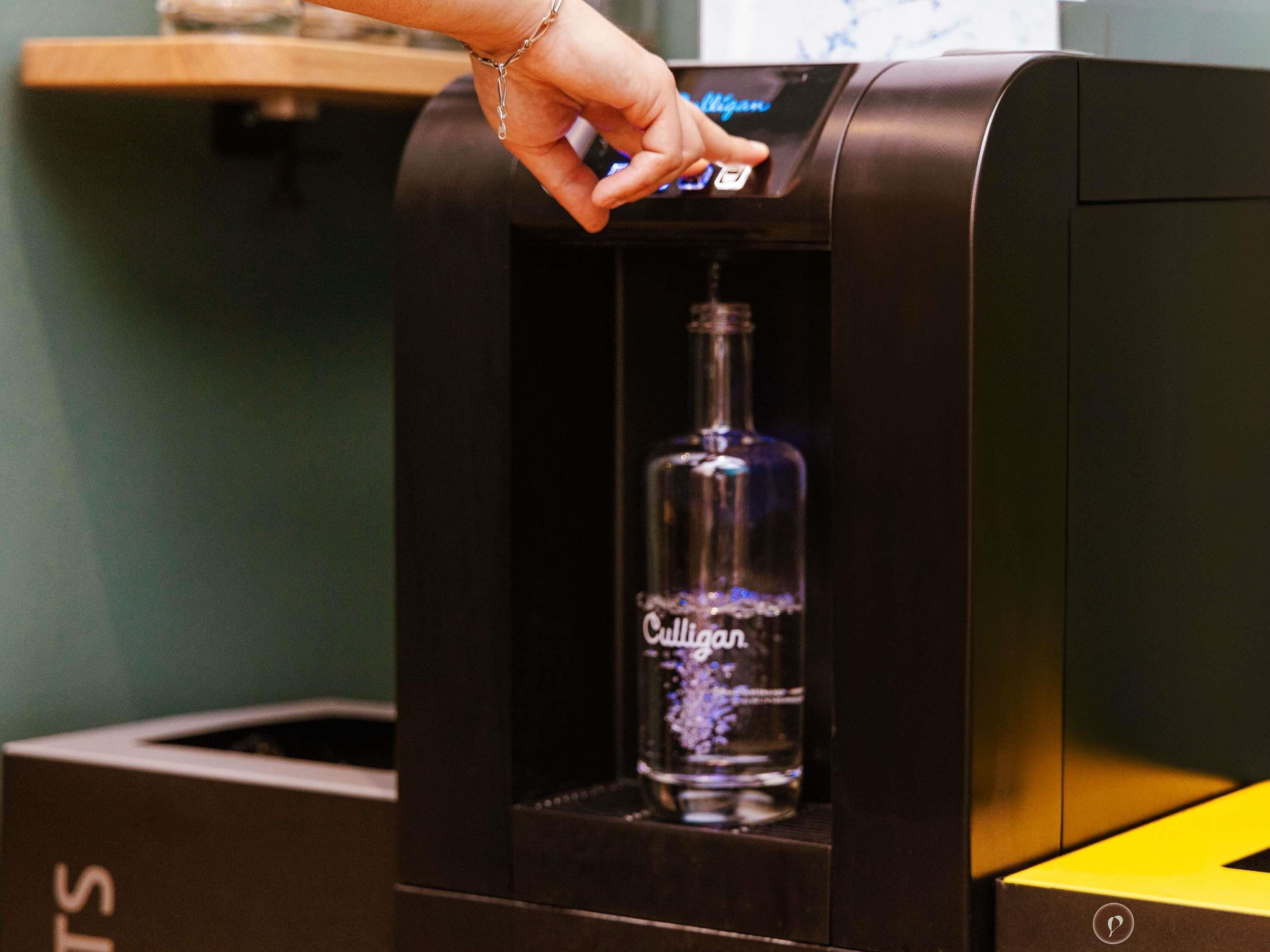 Uma mão interagindo com uma máquina, possivelmente uma máquina de venda automática ou dispensadora, com uma garrafa da marca 'Culligan' no interior.