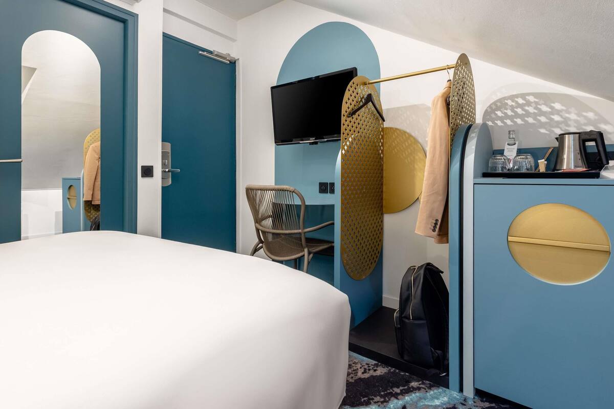 A modern hotel room with a large bed, a TV mounted on the wall, a seating area with a chair, and a dresser with a mirror.