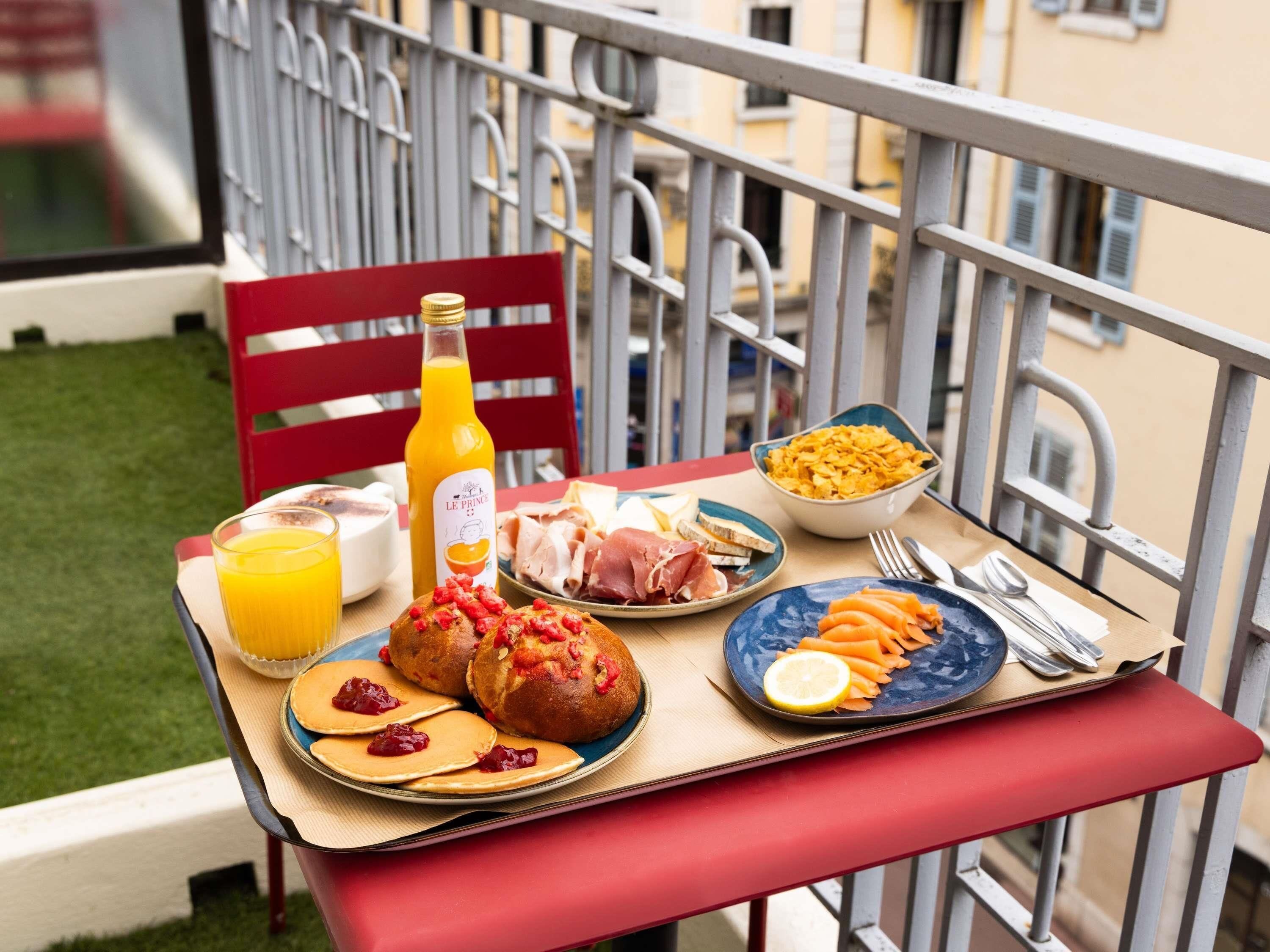 A balcony table with breakfast items including pancakes, sausages, and a bottle of orange juice.