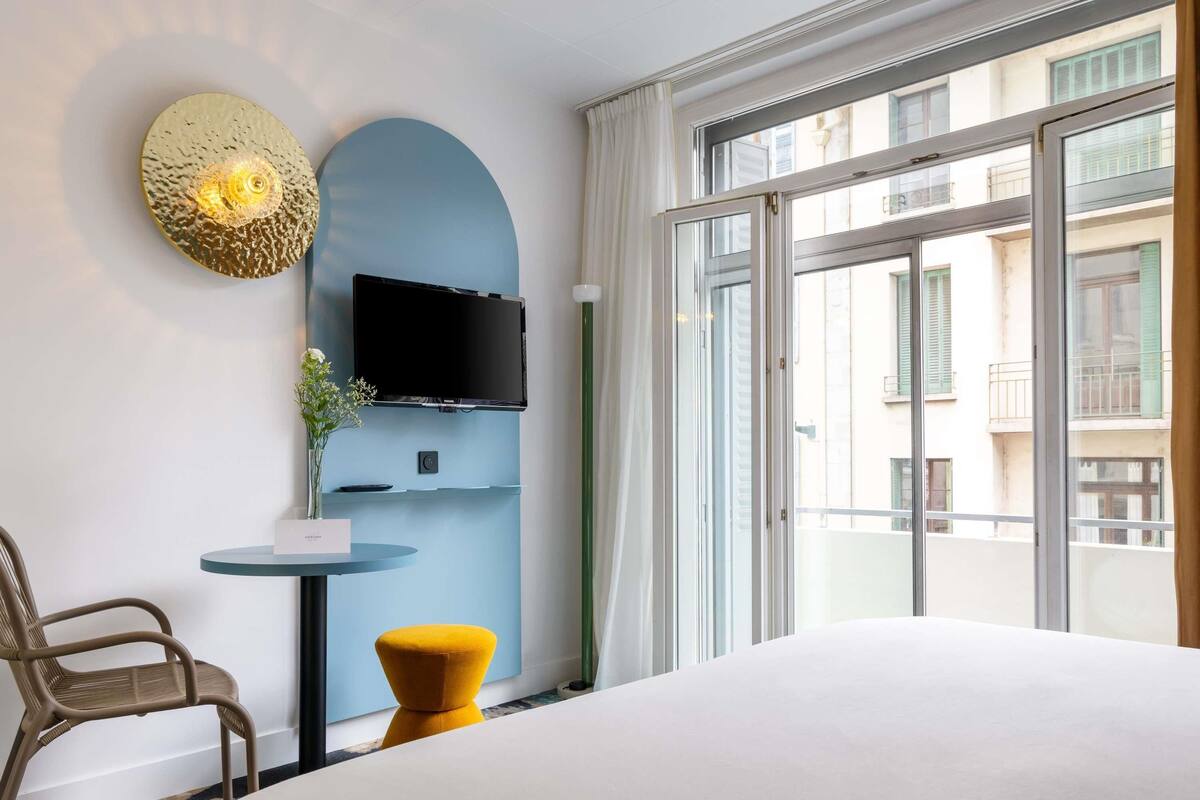 A hotel room with a bed, a TV, a small table with a vase, and a window with a view of buildings.