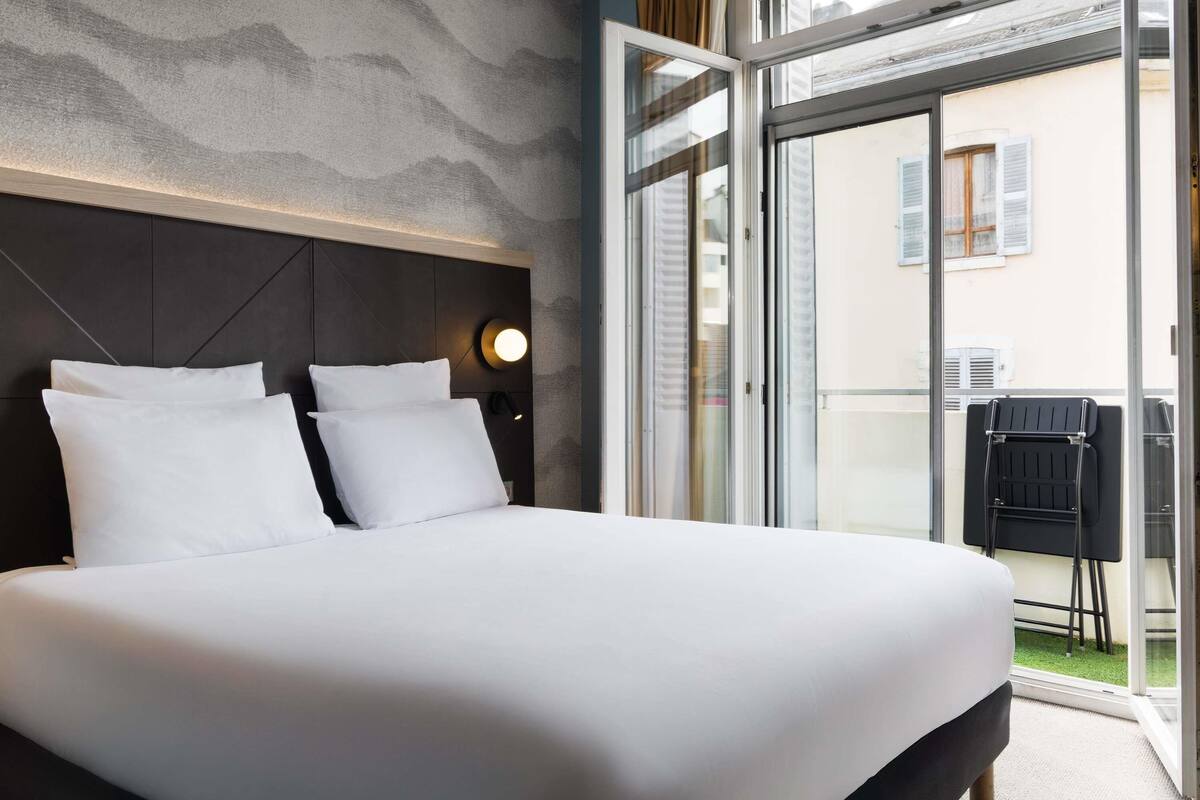 Habitación de hotel con una cama grande, cabecera, luz fijada en la pared y puerta corredera de vidrio que da a un espacio exterior.