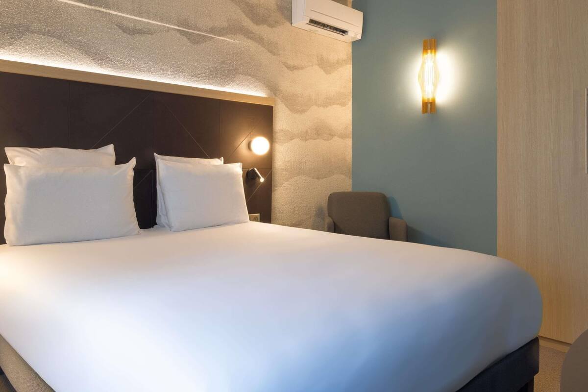 A hotel room with a large bed, a wall-mounted air conditioner, a bedside lamp, and a chair.