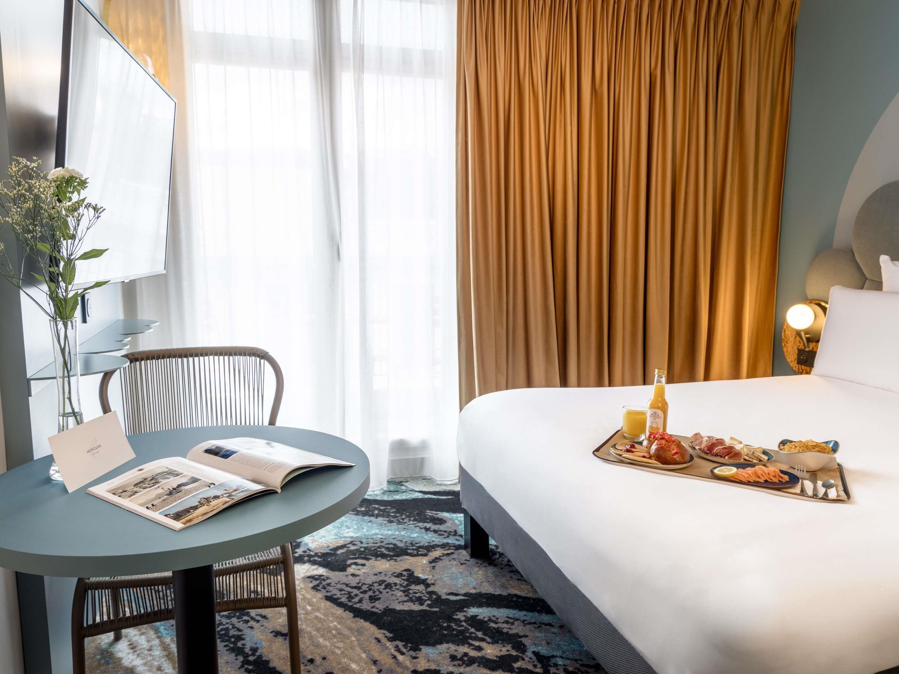 Habitación de hotel con cama, una mesa con bandeja de desayuno, una silla, un espejo y una ventana con cortinas.