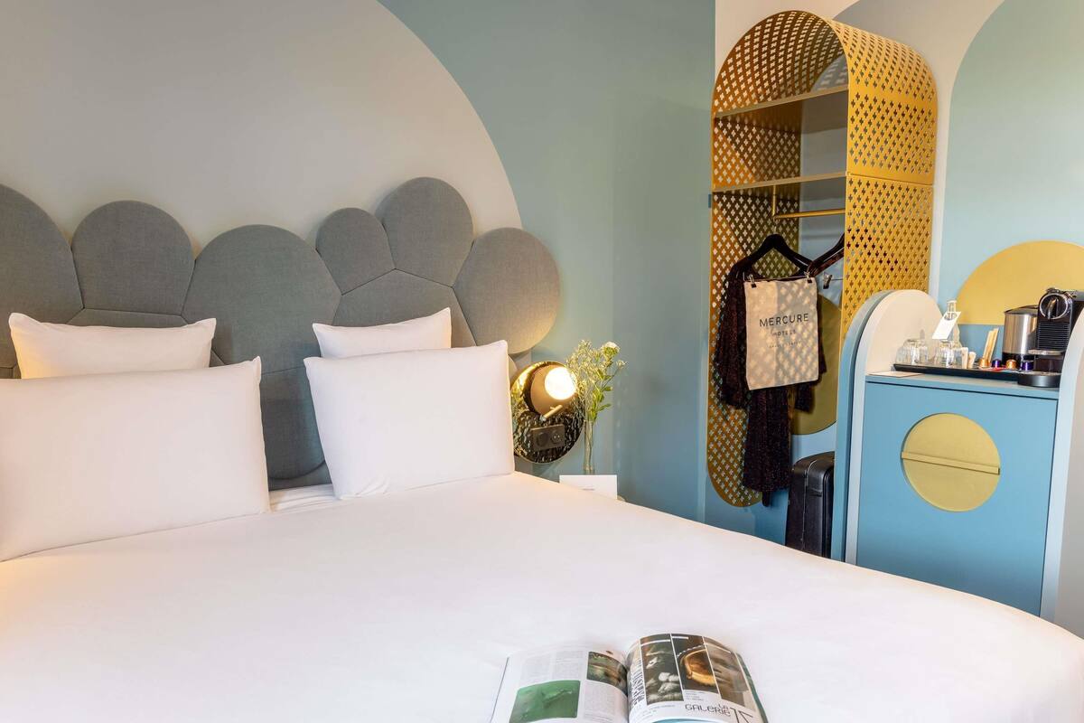A neatly made bed with pillows, a magazine on the bed, a nightstand with a lamp, and a wardrobe with a mirror.