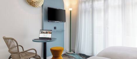 A modern hotel room with a bed, a desk with a laptop, a chair, a TV, and a large window with sheer curtains.