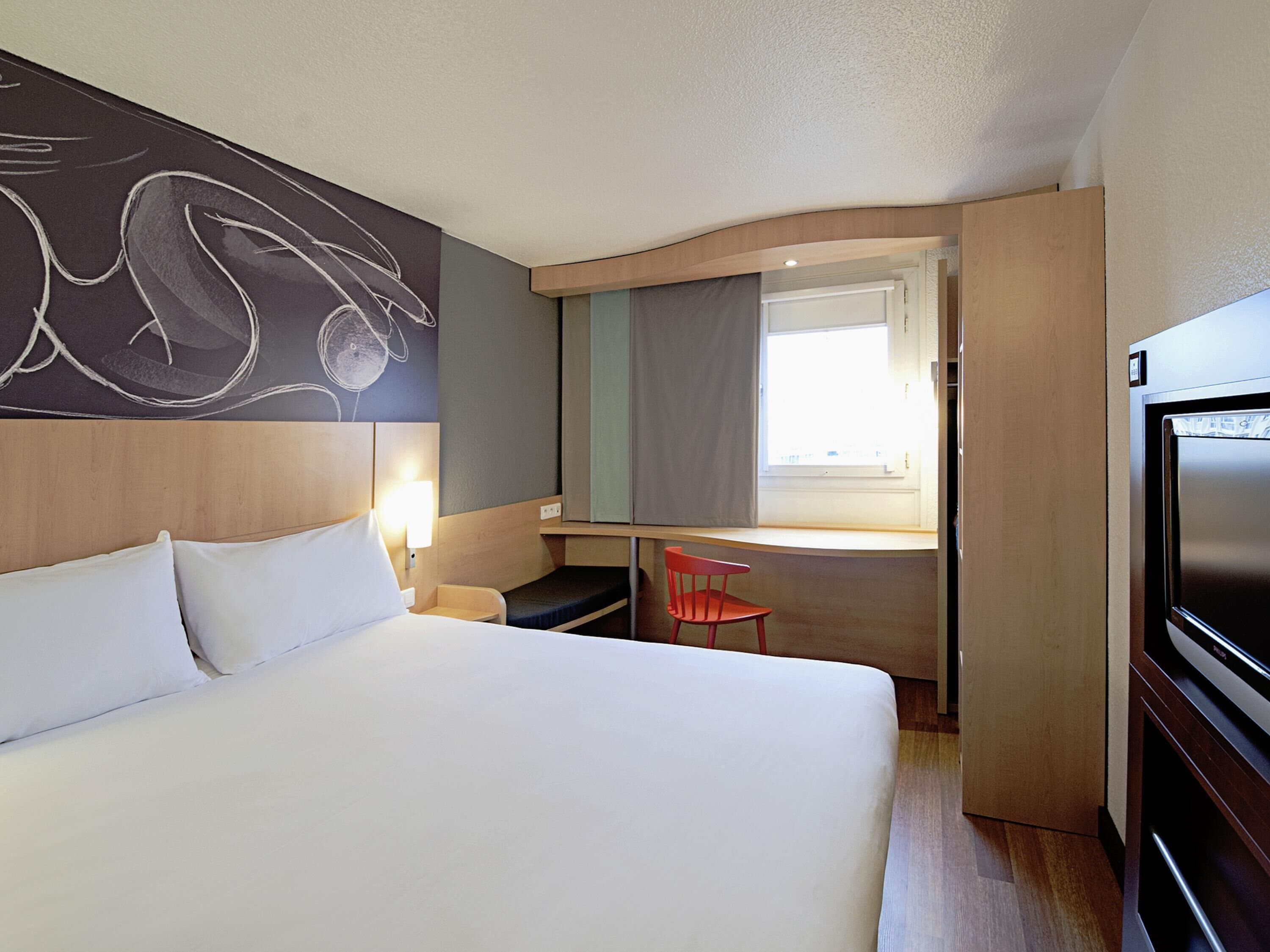A hotel room with a bed, a desk, a chair, a TV, and a window.